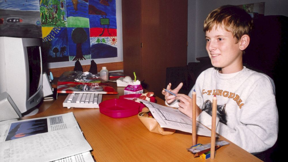 Chłopiec siedzi za biurkiem pełnym bibelotów, na którym stoi też duży biały komputer starego typu. Uśmiecha się szeroko. - grafika artykułu