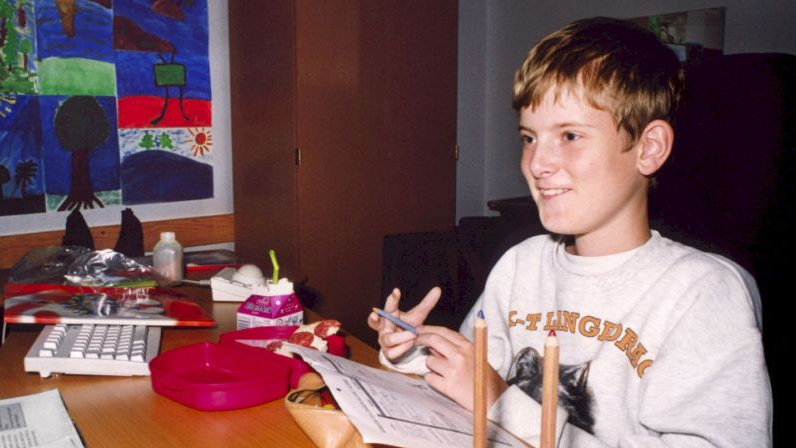 Chłopiec siedzi za biurkiem pełnym bibelotów, na którym stoi też duży biały komputer starego typu. Uśmiecha się szeroko.