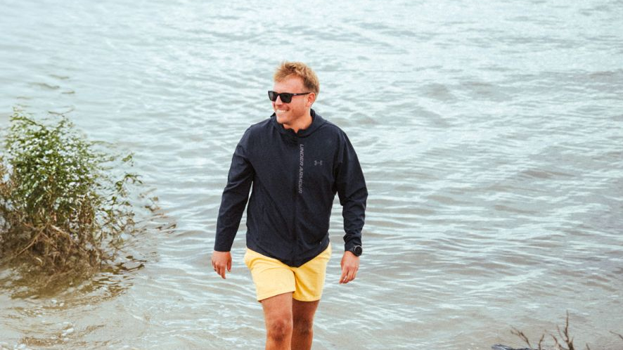 Uśmiechnięty mężczyzna w żółtych szortach i okularach przeciwsłonecznych wychodzi z wody na brzeg.