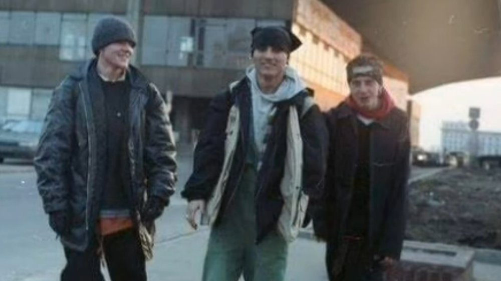 Trzech młodych chłopaków w luźnych ubraniach i czapkach stoi na starym blokowisku. Wszyscy się śmieją, czują się ze sobą swobodnie.