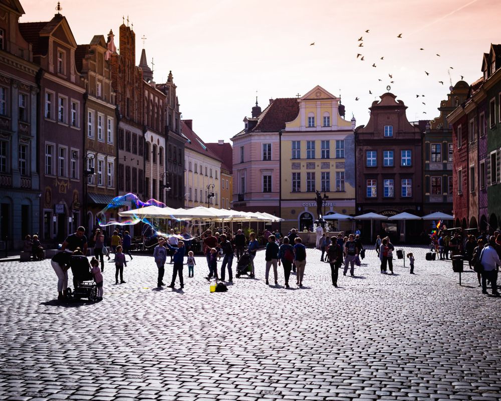 Ludzie spacerujący po Starym Rynku w Poznaniu. Kolorowe fasady kamienic. Ktoś puszcza duże bańki mydlane. - grafika artykułu