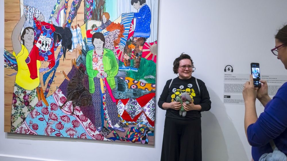 Jedna ze zwiedzających trzyma w rękach małą przytulankę zrobioną ze skrawków. Pozuje obok dużego bardzo kolorowego obrazu, przedstawiającego kobiety wytwarzające ubrania.