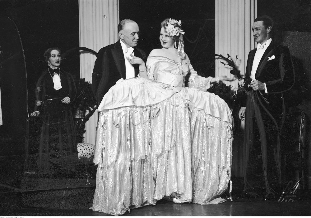 Dwie kobiety i dwóch mężczyzn na scenie. Kobieta w środku kadru ma na sobie białą, rozłożystę suknię balową. - grafika artykułu