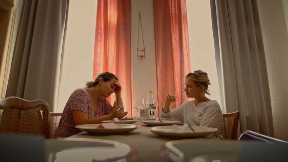 Dwie młode kobiety siedzą przy stole w jasnym pokoju, rozmawiają.