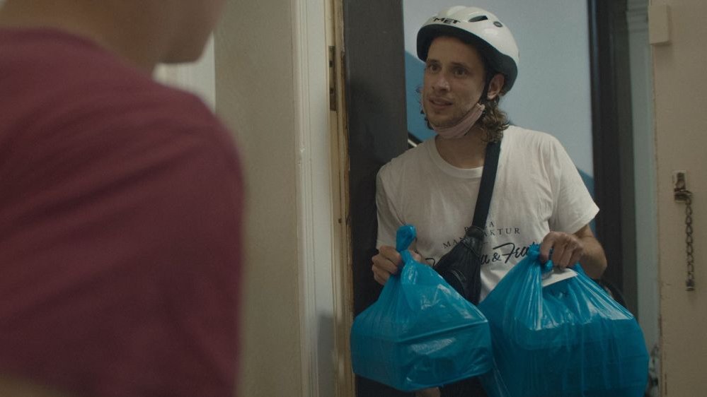 Chłopak w kasku rowerowym stoi w drzwiach, w obu rękach trzyma niebieskie reklamówki z dostarczonym jedzeniem.