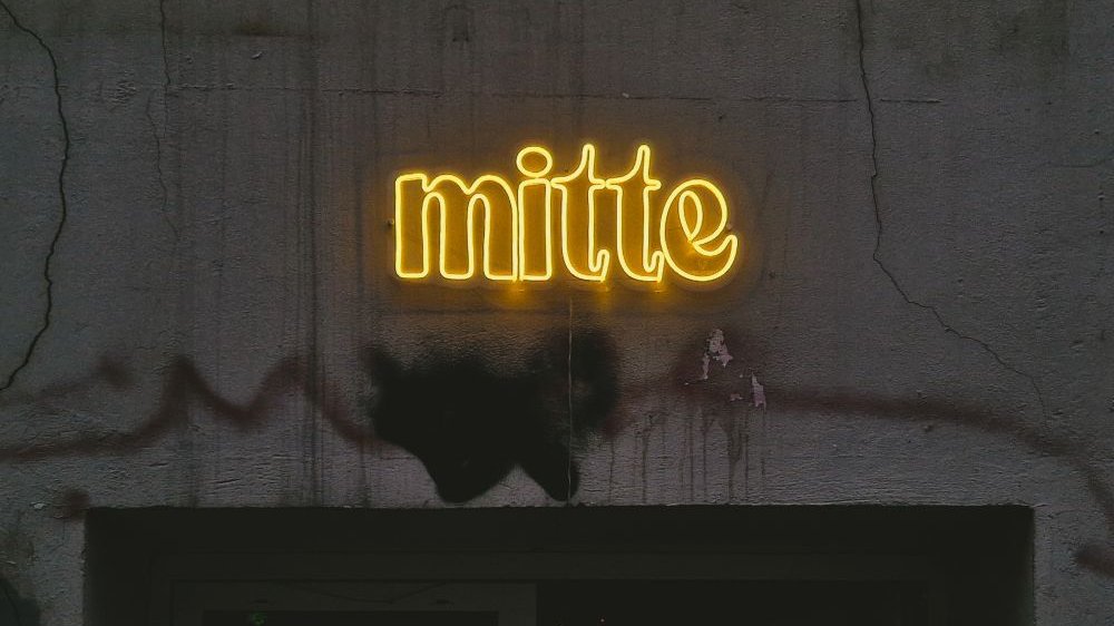 Jaskrawożółty neon ze słowem "mitte" na szarej ścianie. Litery świecą się na obrzeżach, w środku są puste.