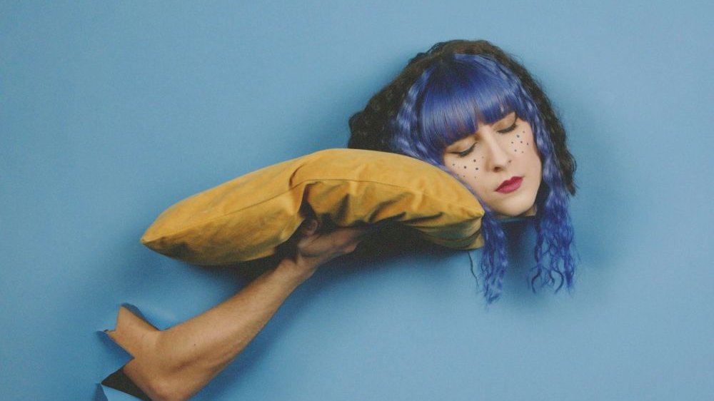 Kolażowe zdjęcie dziewczyny w niebieskich włosach przykładającej głowę do trzymanej przez rękę kogoś innego żółtej poduszki.