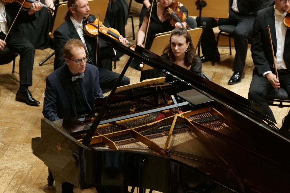 Pianista siedzi przy fortepianie, ma na sobie czarny garnitur, czarną koszulę i szarą muszkę. Patrzy w skupieniu na nuty, obok niego zasiada młoda kobieta, która będzie przewracać stronę za stroną. - grafika artykułu