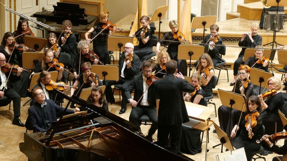 Dyrygent, zwrócony plecami, dyryguje orkiestrze, widzimy kilka rzędów instrumentalistów smyczkowych, zaś po lewej stronie siedzi pianista.