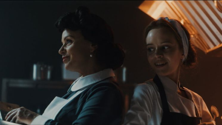 Dwie kobiety w roboczych uniformach siedzą obok siebie, jedna ma rozmarzone spojrzenie, druga, główna bohaterka, zerka w jej kierunku z uśmiechem.