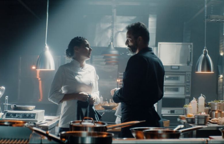 Dwójka bohaterów, kobieta i mężczyzna, stoją naprzeciw siebie w dużej, restauracyjnej kuchni. Ona jest ubrana na biało, on na czarno, patrzą na siebie. - grafika artykułu
