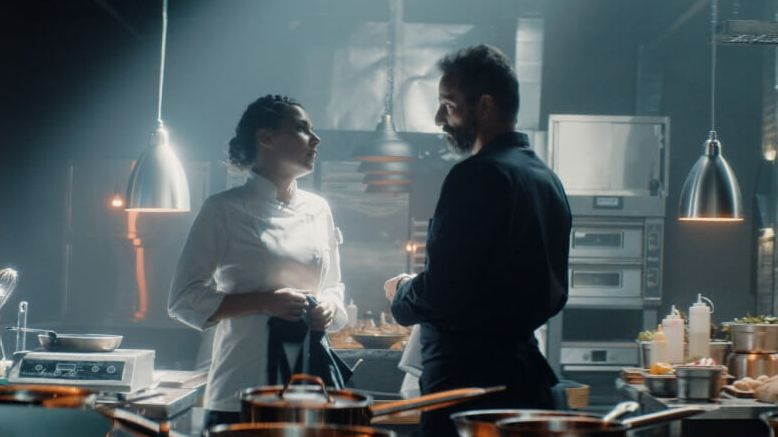 Dwójka bohaterów, kobieta i mężczyzna, stoją naprzeciw siebie w dużej, restauracyjnej kuchni. Ona jest ubrana na biało, on na czarno, patrzą na siebie.