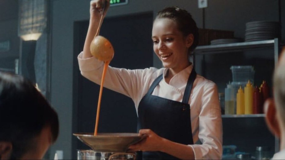 Główna bohaterka w uniformie i fartuchu przyrządza deser, patrzy z radością i apetytem na łyżkę, z której przelewa się do talerza pomarańczowy krem.