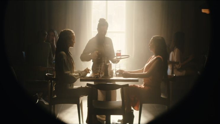 Oświetlone słońcem wnetrze restauracji, przy jednym ze stolików siedzą dwie kobiety, wokół niech krząta się kelnerka, podając napoje.