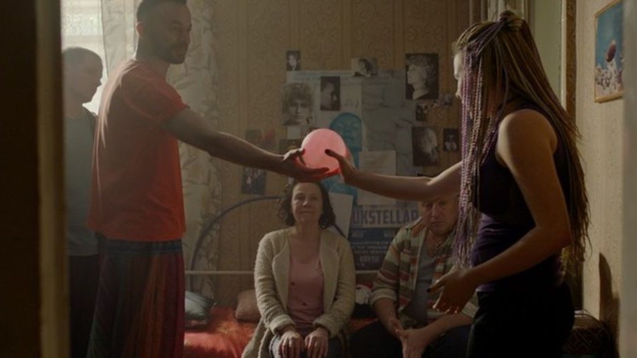 Grupa ludzi gra w grę w pokoju przekazując sobie z ręki do ręki różową piłkę. Na ścianach wiszą plakaty i zdjęcia różnej wielkości.