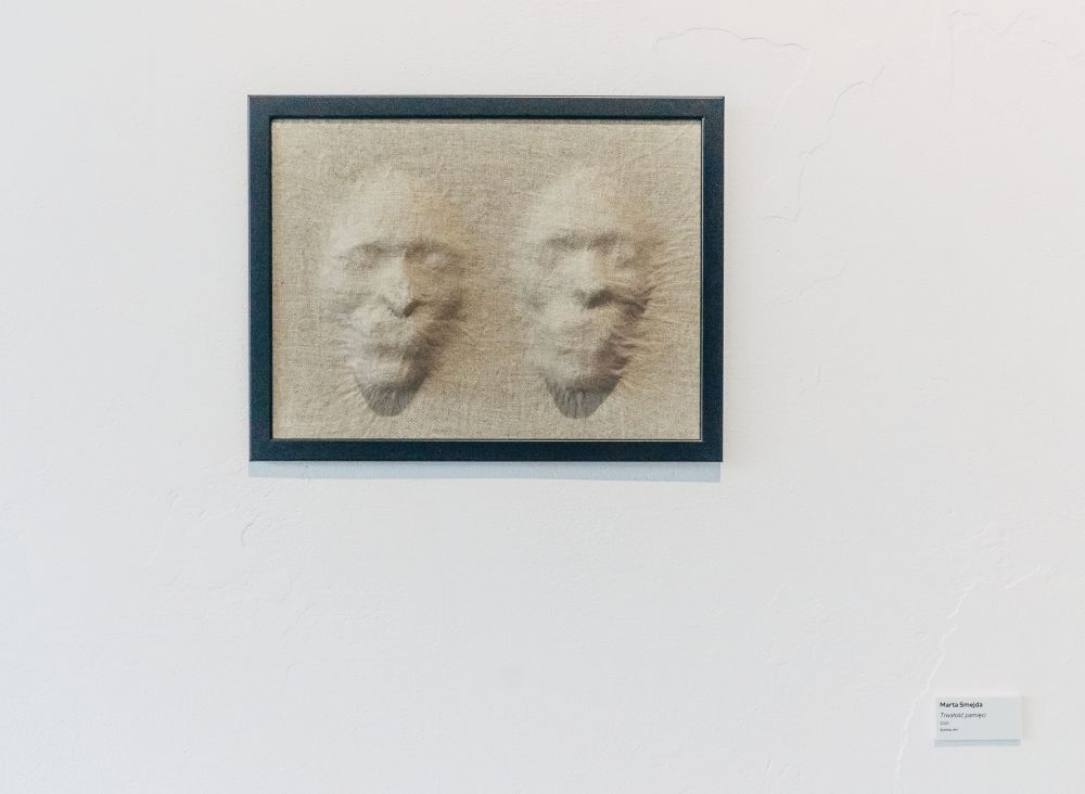 Trójwymiarowy obraz wiszący na ścianie. Przedstawia dwie ludzkie twarze, jakby wypchnięte przez beżową, przypominającą len tkaninę. - grafika artykułu