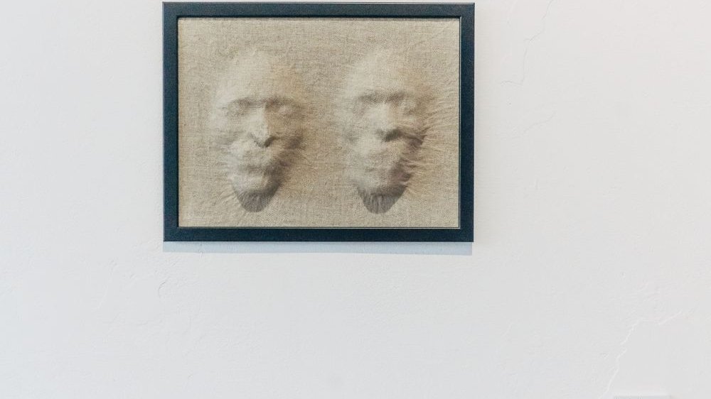 Trójwymiarowy obraz wiszący na ścianie. Przedstawia dwie ludzkie twarze, jakby wypchnięte przez beżową, przypominającą len tkaninę.