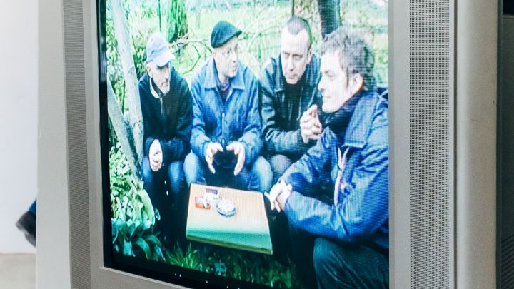 Telewizor starego typu. Na ekranie wyświetla się film z czterema mężczyznami siedzacymi przy małym stoliku w plenerze.
