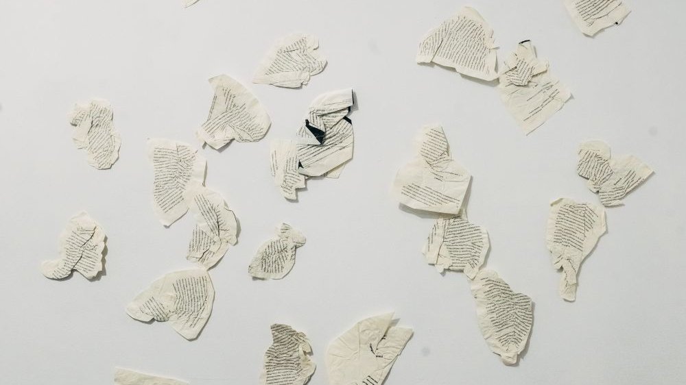 Zgniecione kartki wyrwane najprawdopodobniej z książki i przyklejone do białej ściany galerii.