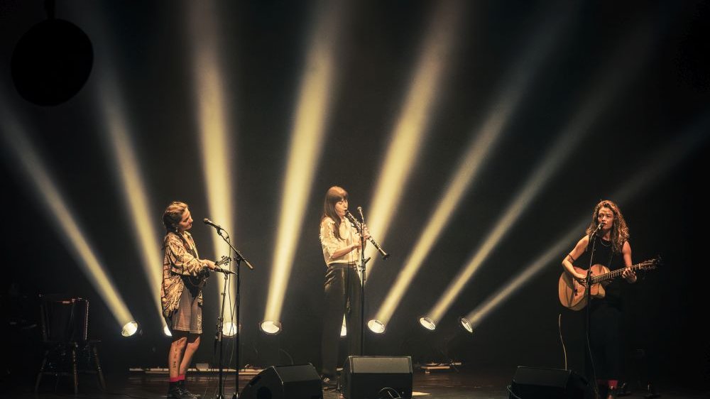 Trzy młode kobiety na scenie. Grają na instrumentach, jedna z nich śpiewa do mikrofonu. Za nimi reflektory światła skierowanego w niebo.