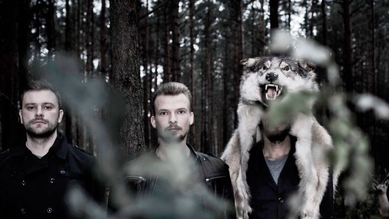 Trzech artystów stoi obok siebie, jeden z nich ma na sobie maskę z wilczym łbem. Za nimi las pełen drzew. Wrażenie jest mroczne, oniryczne, baśniowe.