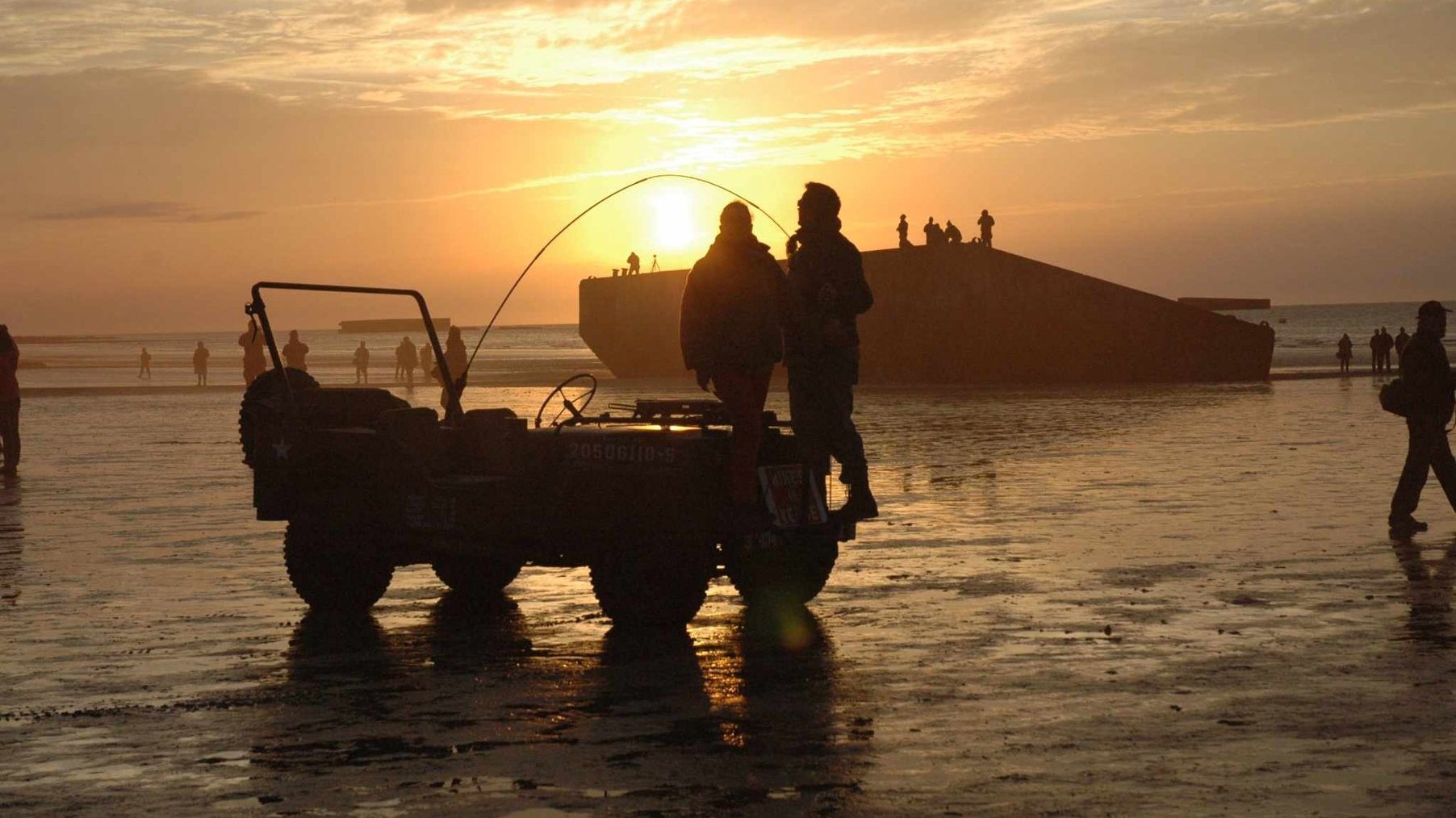 Plaża o zachodzie słońca, na pierwszym tle wojskowy jeep z ludźmi stojącymi na zderzaku, w tle morze.