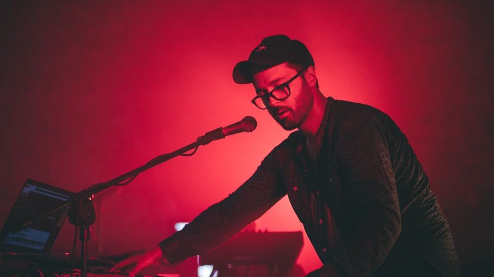 Młody mężczyzna w okularach i czapce z daszkiem gra na klawiszach, przed twarzą ma też mikrofon. W tle czerwone, ciepłe światło.