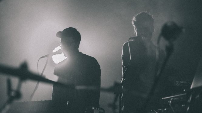 Czarno-białe zdjęcie dwóch muzyków na scenie. Śpiewają do mikrofonów, otacza ich gęsty dym.