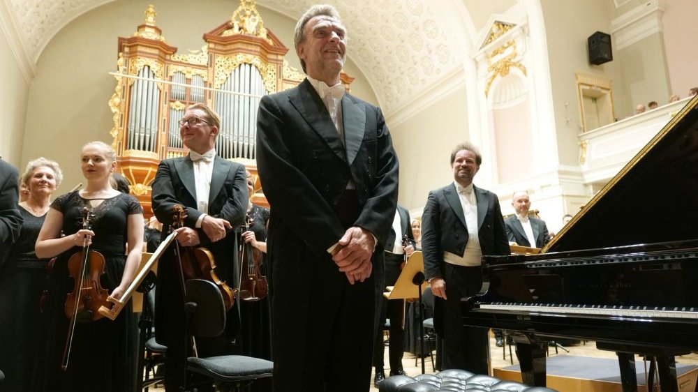 Elegancko ubrani członkowie orkiestry stoją na scenie trzymając instrumenty. Po prawej czarny fortepian, z tyłu złocone organy.