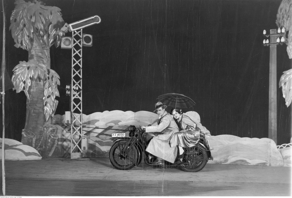 Para na motocyklu w scenicznej scenografii skał i drzew. Kobieta trzyma parasol, pada deszcz. - grafika artykułu