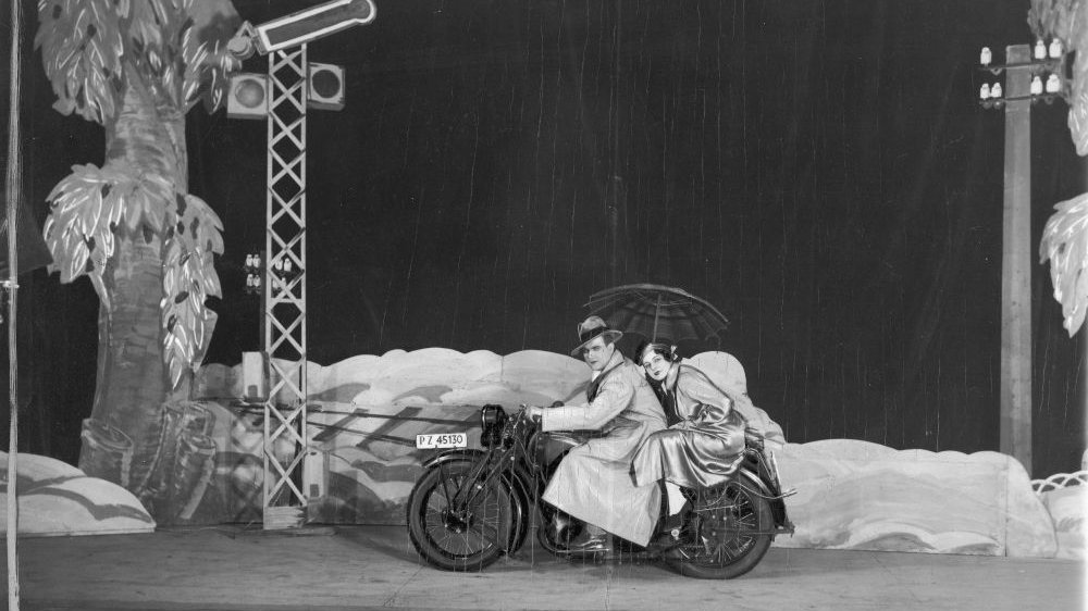 Para na motocyklu w scenicznej scenografii skał i drzew. Kobieta trzyma parasol, pada deszcz.