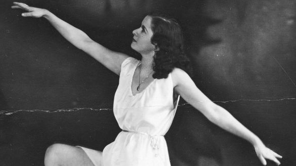 Młoda kobieta w białej sukience wykonuje ruchy baletowe rozkładając szeroko ramiona i unosząc w górę prawą nogę.