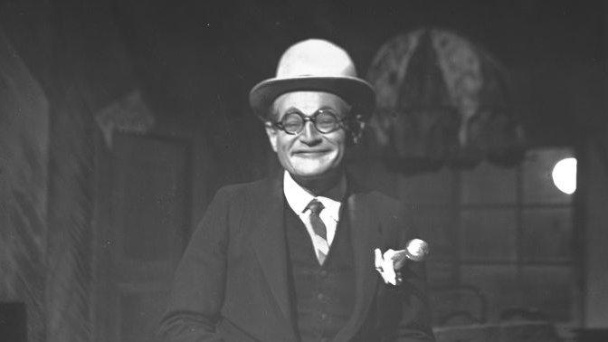 Uśmiechnięty aktor w garniturze, meloniku, okularach i z aktówką występuje na scenie.