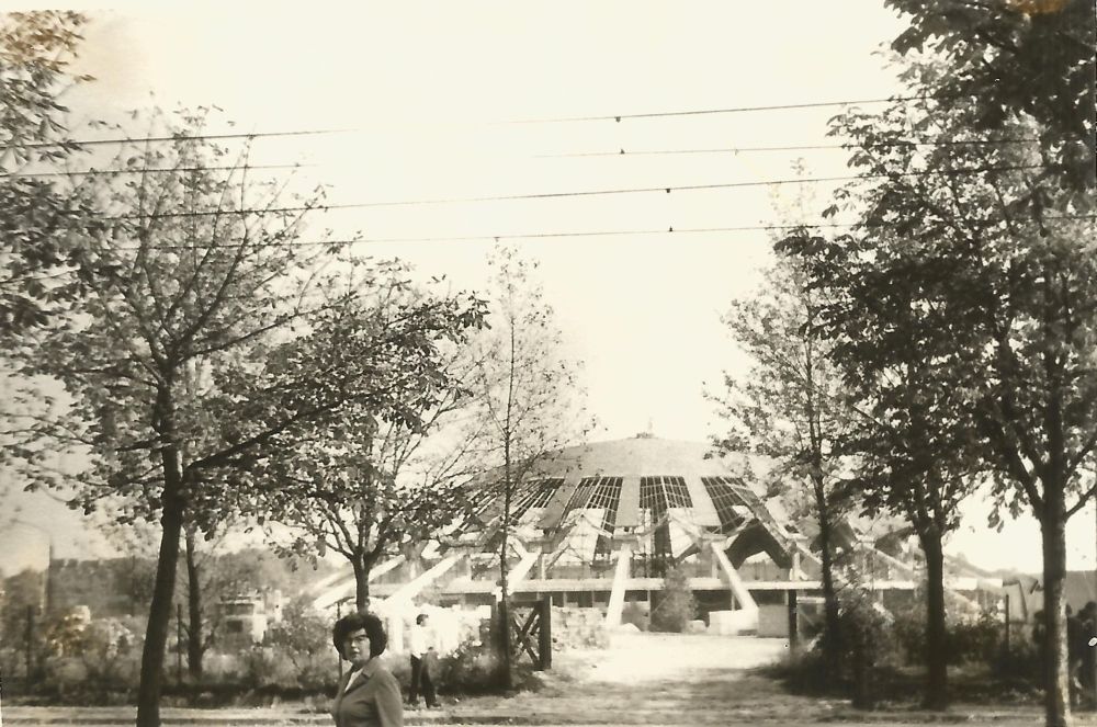 Czarno-białe zdjęcie Areny w budowie. Po prawej i lewej stronie drzewa, z przodu kobieta w okularach. - grafika artykułu