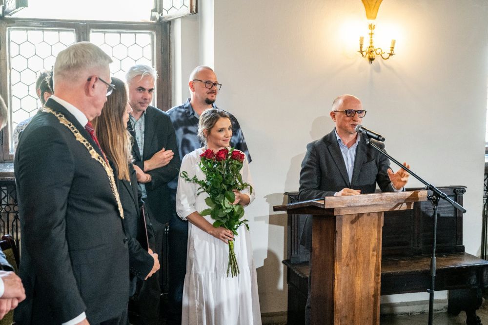 Grupa ludzi odbiera nagrodę w zabytkowym ratuszu. Z mównicy przemawia mężczyzna, kobieta obok niego trzyma bukiet róż. Po lewej stoi prezydent miasta w złotym łańcuchu na szyi. - grafika artykułu