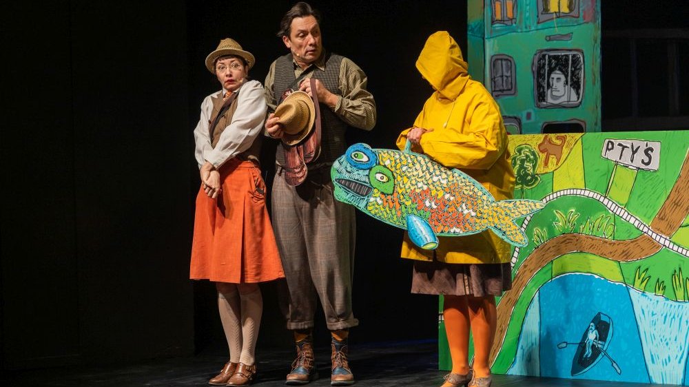 Bohaterowie, Dora Dynks i Waldemar Wihajster, stoją wyraźnie wstrząśnięci obok postaci w żółtej kurtce przeciwdeszczowej i pomarańczowych rajstopach, któa niesie wielką, wyszczerzoną, rysunkową rybę.