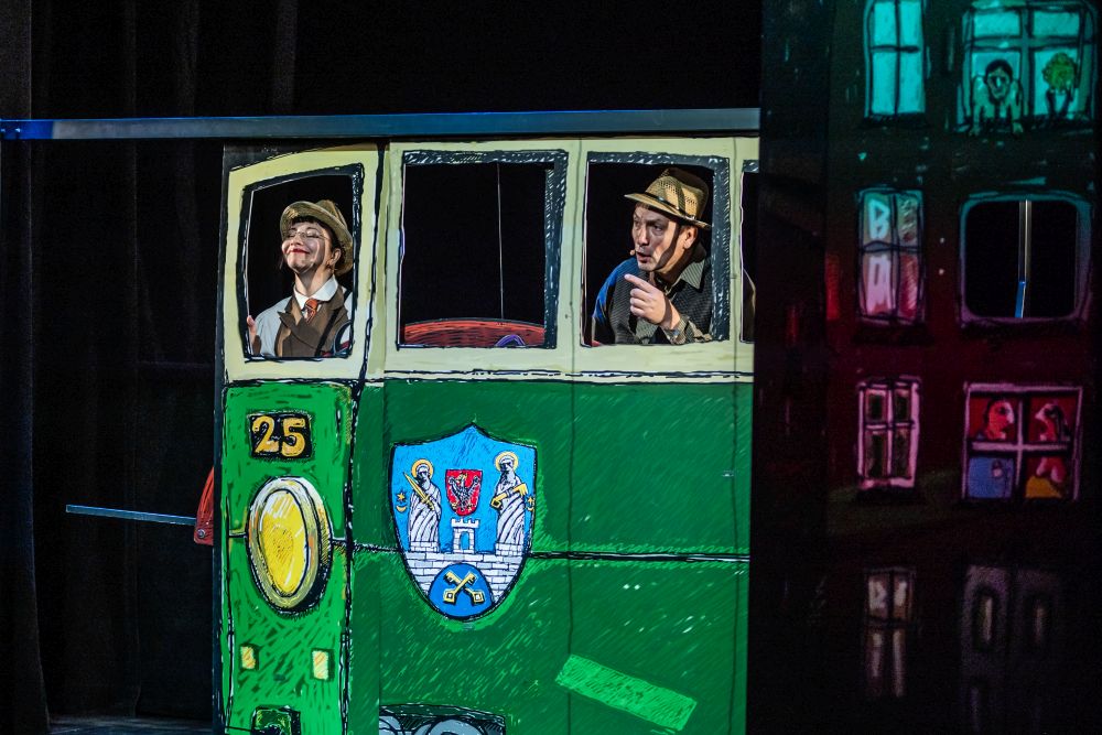 Zielony tramwaj o numerze 25, a właściwie jego teatralna, malowana farbami replika. Jadą nim bohaterowie spektaklu, Dynks uśmiecha się radośnie, Wihajster wygląda na czujnego i wskazuje coś palcem. - grafika artykułu