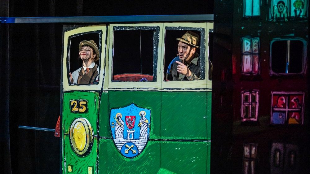 Zielony tramwaj o numerze 25, a właściwie jego teatralna, malowana farbami replika. Jadą nim bohaterowie spektaklu, Dynks uśmiecha się radośnie, Wihajster wygląda na czujnego i wskazuje coś palcem.