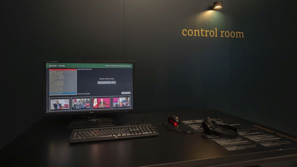 Biurko z monitorem komputera, klawiaturą, myszką i słuchawkami. Na czarnej ścianie napis "control room".