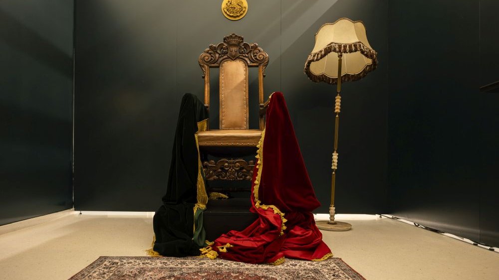 Bogato rzeźbione krzesło stoi obok tureckiego dywanu i staromodnej lampy z kloszem. Na obu jego poręczach wiszą lejące materiały, jeden w kolorze czarnym, drugi w purpurowym.