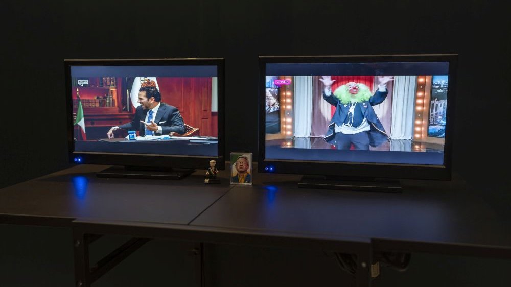 Dwa monitory stoją na czarnym blacie. Na jednym z nich film ze śmiejącym się mężczyzną, prawdopodobnie politykiem, na drugim klaun podczas występu.