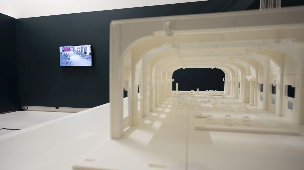 Po prawej widok na makietę budynku wykonaną z białego tworzywa, po lewej monitor z wyświetlonym filmem wiszący na ścianie.