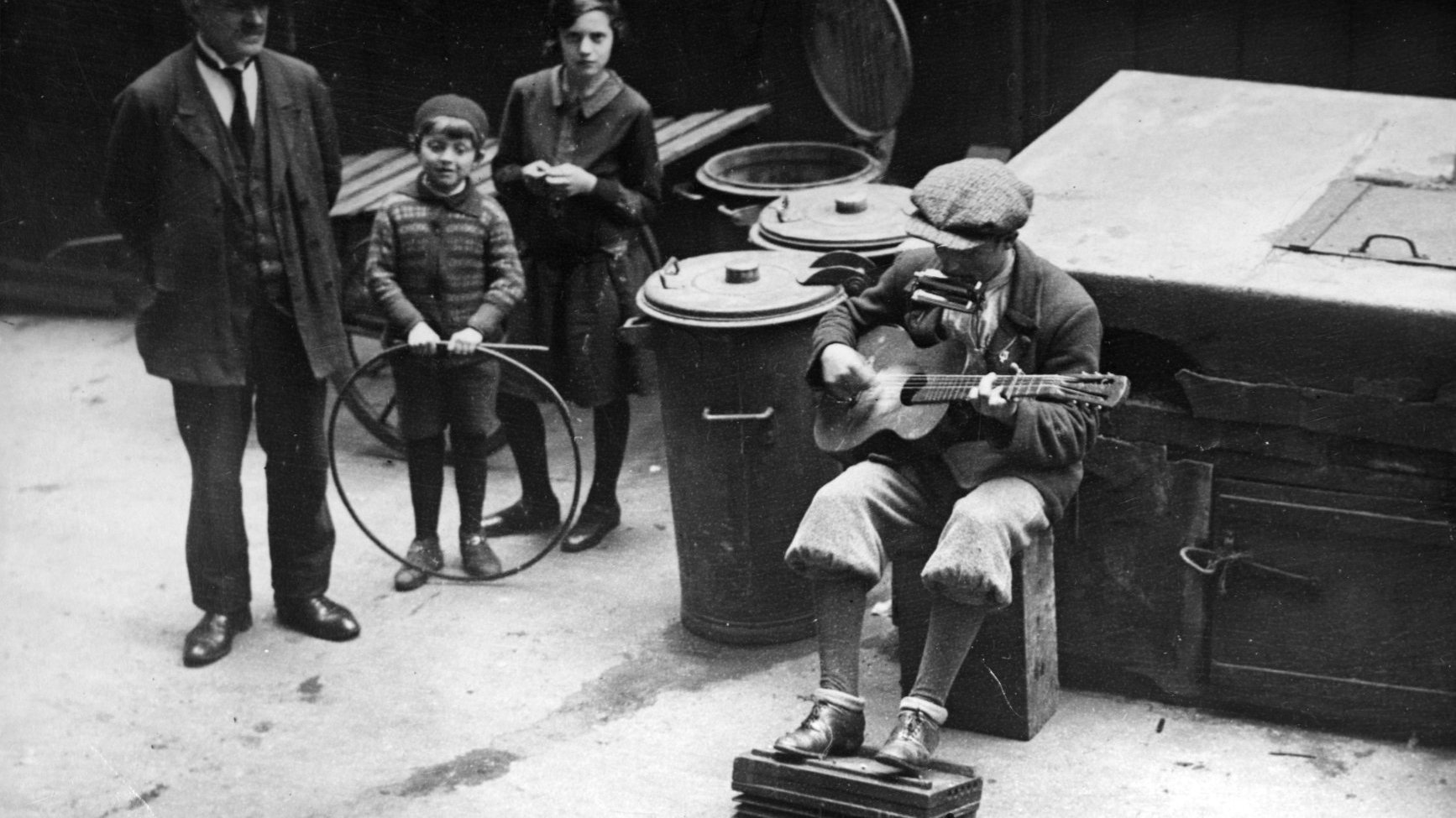 W centrum czarno-białej fotografii jest muzyk z gitarą. Słucha go starszy meżczyna, młoda kobiea i dziecko. Muzyk siedzi obok metalowych kubłów na śmieci.