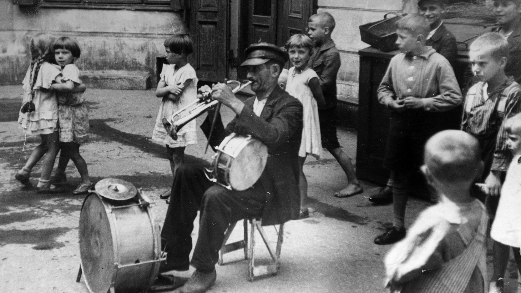 W centrum czarno-białej fotografii jest muzyk z parkusją i trąbką. Słucha go grupa dzieci, dwie dziewczynki tańczą.