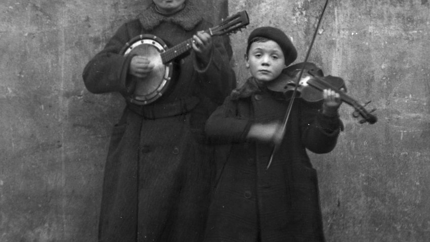 W centrum czarno-białej fotografii na tle zimnego muru jest dwoje dzieci w zimocyh płaszczach. Niższe gra na skrzypach, a wyższe na intrumencie przypomniającym bałałajkę. Dzieci są przygnębione.