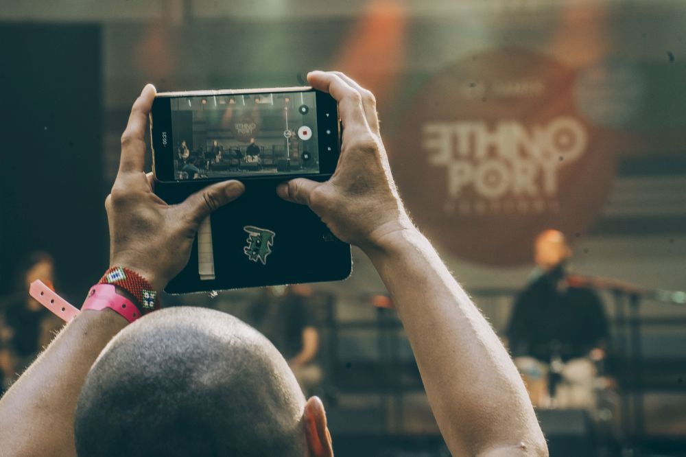 Mężczyzna jest w trakcie robienia zdjęcia - widzimy jego wystawione ręce i obiektyw telefonu. Przed nim scena z logo "Ethno port", na scenie występuje muzyk. - grafika artykułu