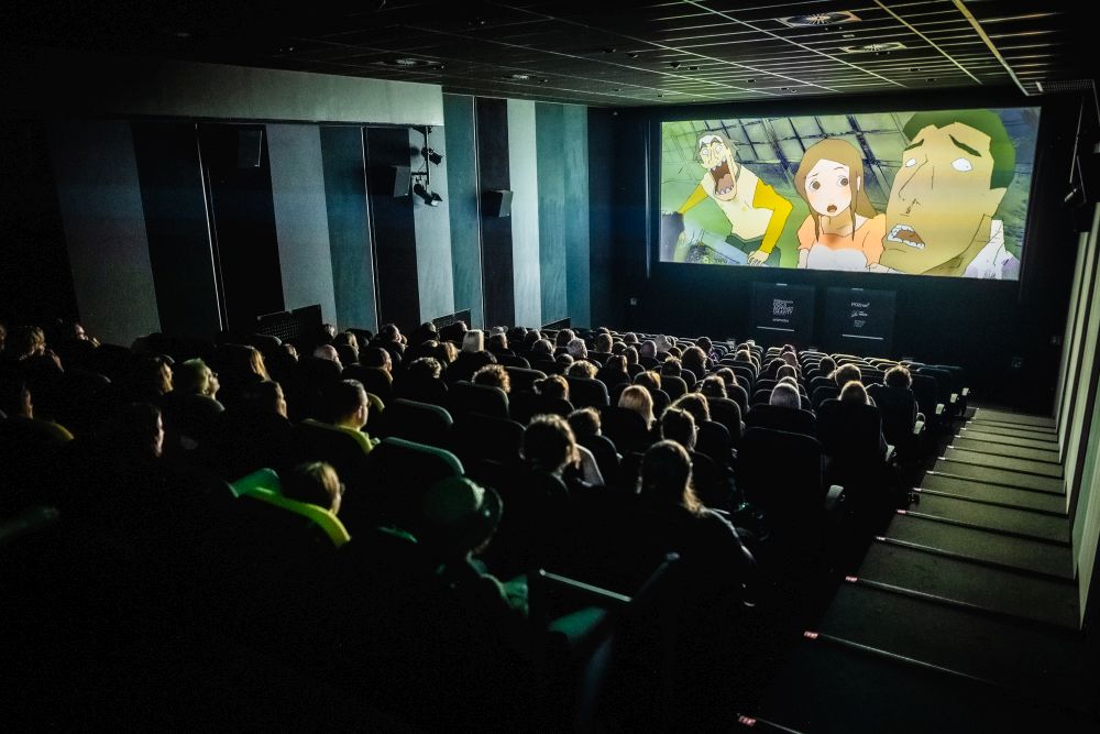 Publiczność festiwalowa ogląda film animowany w kinie Muza. W sali jest ciemno, głowy widzów oświetla jedynie światło z ekranu. - grafika artykułu