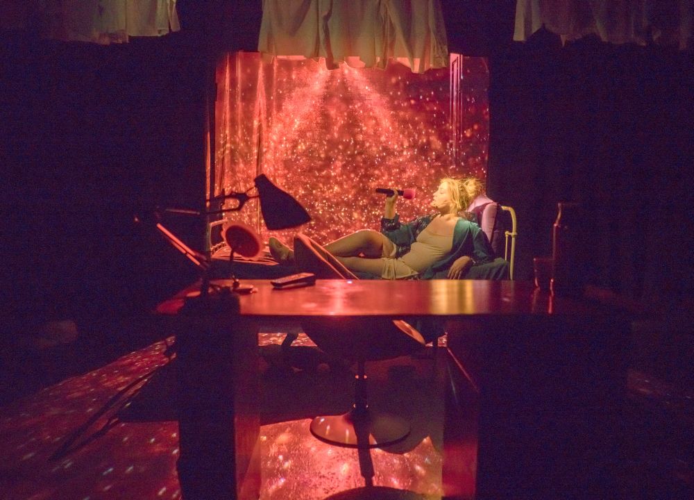 Jasnowłosa kobieta śpiewa przez różowy półleżąc na kanapie. Jest oświetlona czerwonym, jaskrawym światłem, które wygląda magicznie, jak otaczający ją pyłek. - grafika artykułu