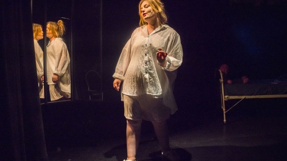 Bohaterka stoi w ciemności przed lustrem, ma na sobie białą koszulę, spódnicę i bambosze w kształcie głów misia.