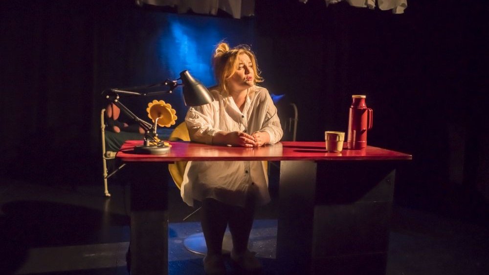 Bohaterka siedzi zamyślona przy czerwonym biurku, na którym stoi czarna lampka, lusterko w kształacie żółtego kwiatka (słonecznika) oraz termos i kubek.